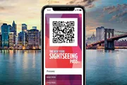 A smartphone displays a New York Sightseeing Pass with a QR code on the screen, superimposed on a background featuring a city skyline at dusk.