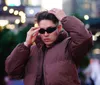 A person is adjusting their sunglasses and hair while wearing a brown jacket in an urban outdoor setting