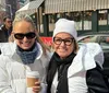 Two smiling women in winter clothing are holding coffee cups on a sunny city street