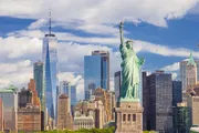 The image features the Statue of Liberty in the foreground with the New York City skyline, including One World Trade Center, in the background under a partly cloudy sky.