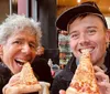 Two people are smiling and holding up large slices of pizza to their mouths as if taking a bite in a casual dining setting