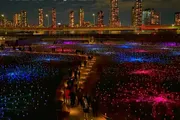 The image shows a vibrant field of multicolored lights in the foreground with visitors walking through, set against a backdrop of a city skyline at night.