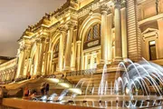 The image shows an illuminated classical building at night with a grand staircase and an elegant fountain in the foreground, likely a museum or a similar cultural institution.