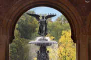 The image shows a statue of an angel with outstretched wings atop a fountain, framed by an archway, with a backdrop of autumnal trees.