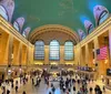 The photo shows the bustling interior of Grand Central Terminal with people milling about under the expansive arched windows and a large American flag hanging prominently