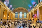 The photo shows the bustling interior of Grand Central Terminal, with people milling about under the expansive arched windows and a large American flag hanging prominently.