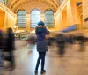 The photo shows the bustling interior of Grand Central Terminal with people milling about under the expansive arched windows and a large American flag hanging prominently