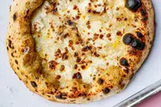 The image shows a freshly baked pizza with a golden-brown crust and melted cheese, speckled with charred spots typical of a wood-fired oven.