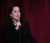 A person is performing stand-up comedy on a stage with a brick wall background and a cityscape sign