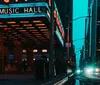 The image showcases an evening city scene with pedestrians and cars illuminated by the retro neon signage of the Music Hall