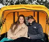 A smiling man and woman are sitting together in a yellow horse-drawn carriage with a snowy backdrop