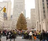 The image shows a bustling crowd of people gathered around a large decorated Christmas tree in an urban setting indicative of a holiday celebration in a city