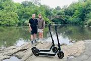 Two individuals and two electric scooters are standing on a rock with a pond and an arched bridge in the background.