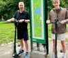 Two individuals and two electric scooters are standing on a rock with a pond and an arched bridge in the background