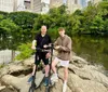 Two individuals and two electric scooters are standing on a rock with a pond and an arched bridge in the background