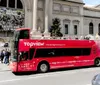 A red double-decker sightseeing bus marked TopView is parked in front of a neoclassical building with people sitting on top and others milling about indicating a bustling city scene likely popular with tourists