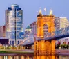 The image captures a lit-up stone bridge with intricate arches spanning a river leading towards a modern city skyline illuminated against the dusk sky