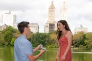 A person is proposing to their partner with a ring by a lake with city buildings in the background.