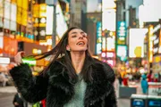 A joyful woman is playfully flipping her hair in a bustling city street illuminated by vibrant neon signs at dusk.