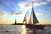 A sailboat is gliding across the water near the Statue of Liberty during a beautiful sunset.