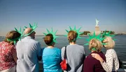 A group of tourists wearing green foam Statue of Liberty crowns gazes at the real Statue of Liberty from a boat.