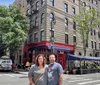 A man and a woman are smiling for a photo on a tree-lined street with a classic urban building in the background featuring a ground-floor restaurant and an outdoor dining area