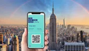 A hand holds up a smartphone displaying a New York Pass ticket with a QR code in front of a panoramic view of New York City.