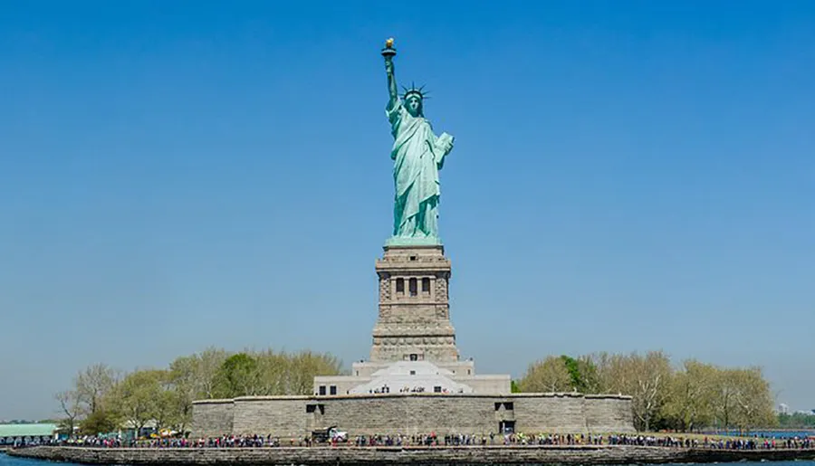The image shows the Statue of Liberty standing tall against a clear blue sky, with numerous visitors visible at its base.