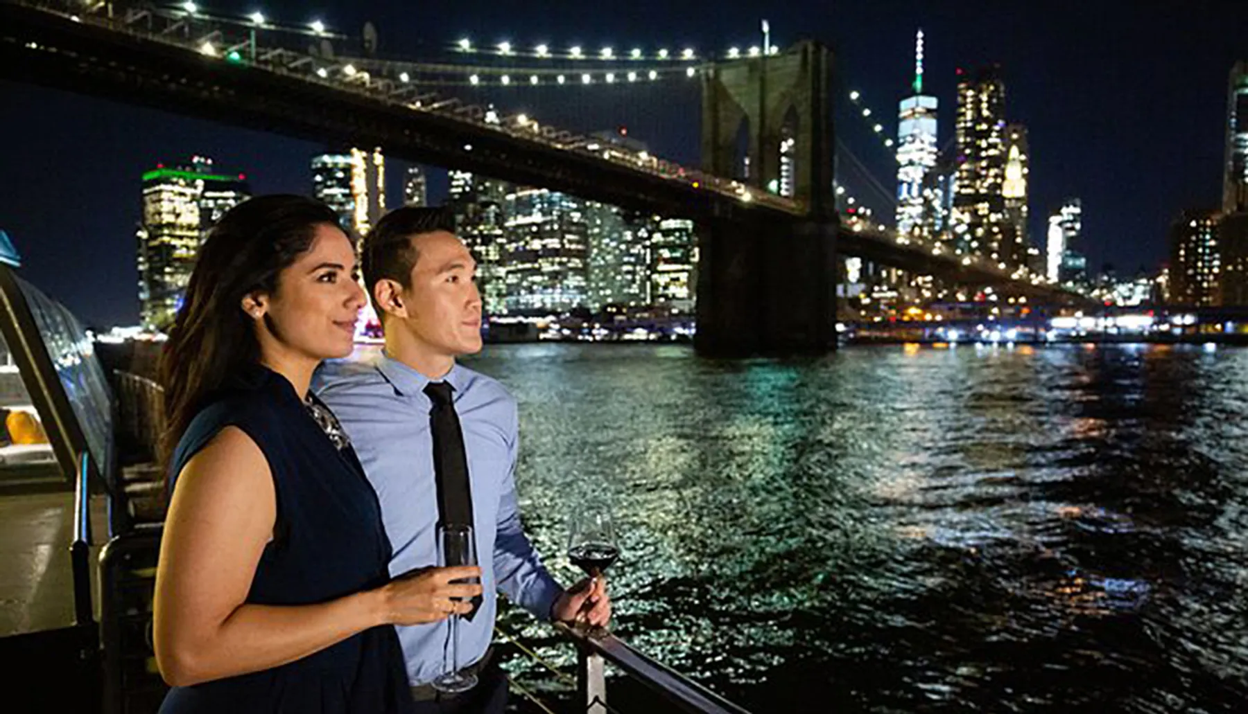 A couple enjoys a nighttime view of a lit-up cityscape with a prominent bridge in the background, while holding glasses of wine.