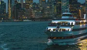 A cruise ship filled with passengers sails in front of a brightly lit city skyline during twilight hours.