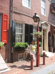 A person is standing on a quaint urban street with brick buildings, flower boxes, and a classic lamp post on a sunny day.