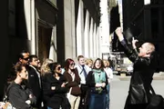A group of people listens attentively as a man in a black jacket animatedly speaks or performs, possibly on a city street or during a walking tour.