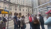 A group of people appear to be engaged in a guided tour on a city street lined with tall buildings, some walking by and others stopping at a food cart.