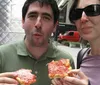 A man and woman are casually standing and eating slices of pizza on a city street