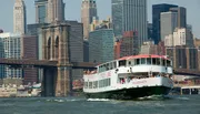 A sightseeing boat cruises near the Brooklyn Bridge with the backdrop of the Manhattan skyline.