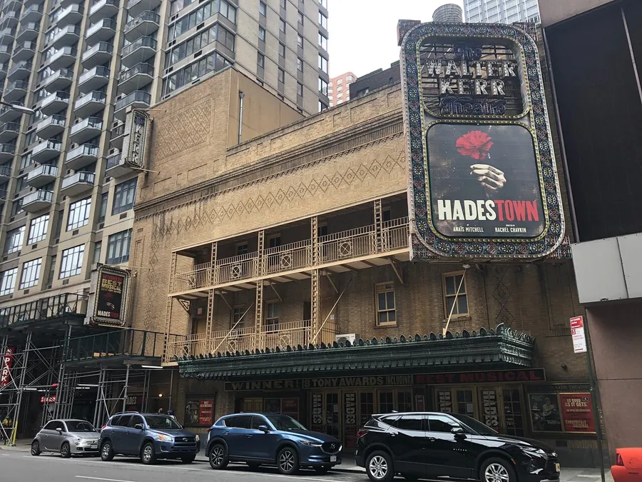 The image shows the exterior of the Walter Kerr Theatre, advertising the musical Hadestown on its marquee, with cars parked in front and a modern building towering in the background.