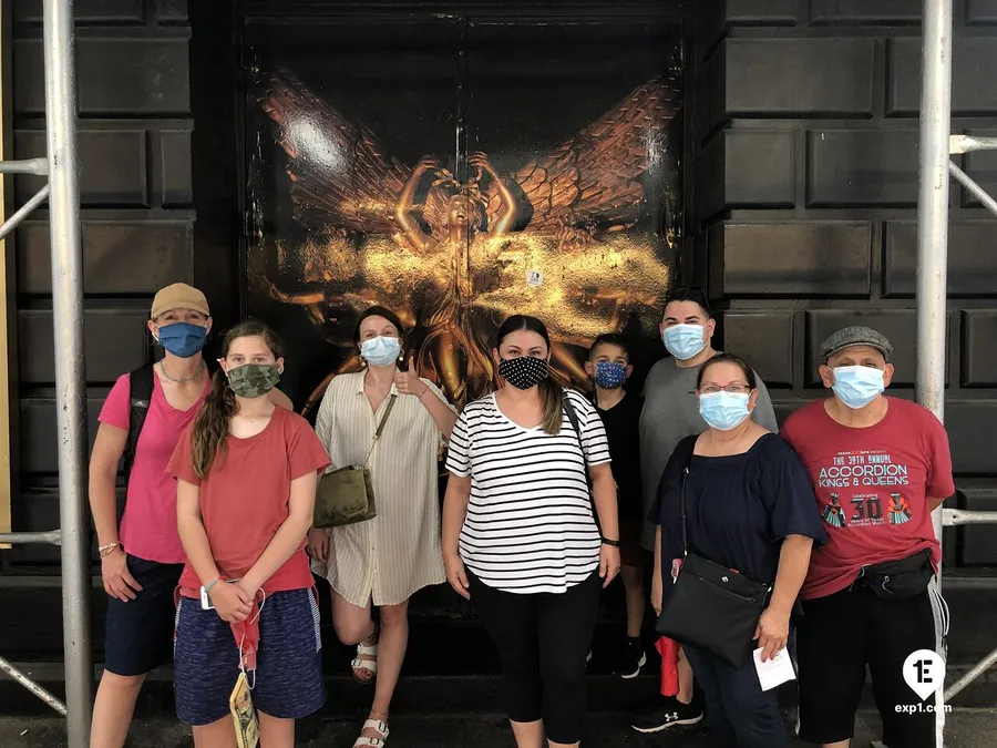 A group of people wearing protective face masks are posing for a photo in front of an ornate door with a striking golden insect design.