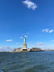 The photo captures the Statue of Liberty standing on Liberty Island against a backdrop of clear blue sky with scattered clouds, viewed from the perspective of the surrounding water.