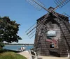 A traditional wooden windmill stands by a clear blue sky with an American flag out front while people enjoy a sunny day by the water