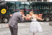 A man bows to a joyful woman in a dress as amazed passengers on a tour bus look on.