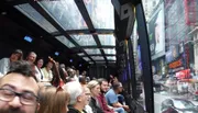 The image captures a group of people on a transparent-roofed sightseeing bus tour enjoying the cityscape, with one person enthusiastically pointing out a feature.