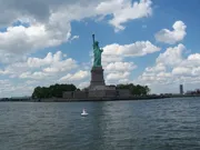 The image shows the Statue of Liberty on its island under a partly cloudy sky, as seen from a distance over the water with a buoy visible in the foreground.