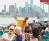 A tour guide in a yellow jacket is speaking into a microphone and pointing something out to a diverse group of attentive passengers on a boat tour with a city skyline in the background