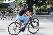 A person wearing a helmet and sunglasses is riding a road bike on a street alongside other cyclists in casual attire.