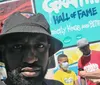 A man in a black hat is taking a selfie with a colorful graffiti wall in the background while several people including one wearing a mask are engaging with each other in the background