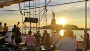 A group of people is enjoying a live music performance on a boat at sunset with the Statue of Liberty in the background.