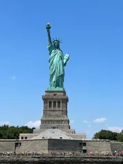 The image shows a full view of the Statue of Liberty against a clear blue sky, with visitors visible at its base on Liberty Island.