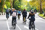 A group of people are cycling along a tree-lined road in what appears to be an urban park setting with autumn foliage.