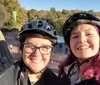 Two people with helmets are smiling for a selfie with a park featuring trees and people in the background suggesting they might be enjoying an outdoor activity like cycling