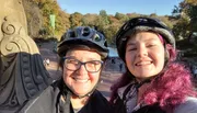 Two people with helmets are smiling for a selfie with a park featuring trees and people in the background, suggesting they might be enjoying an outdoor activity like cycling.